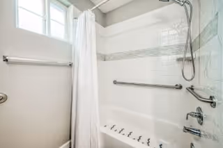 A bathroom featuring a white bathtub with multiple water jets, white tiled walls with a horizontal gray accent strip, a silver showerhead, and grab bars on the wall. A white shower curtain hangs on a rod, and a window above the tub allows natural light into the space.