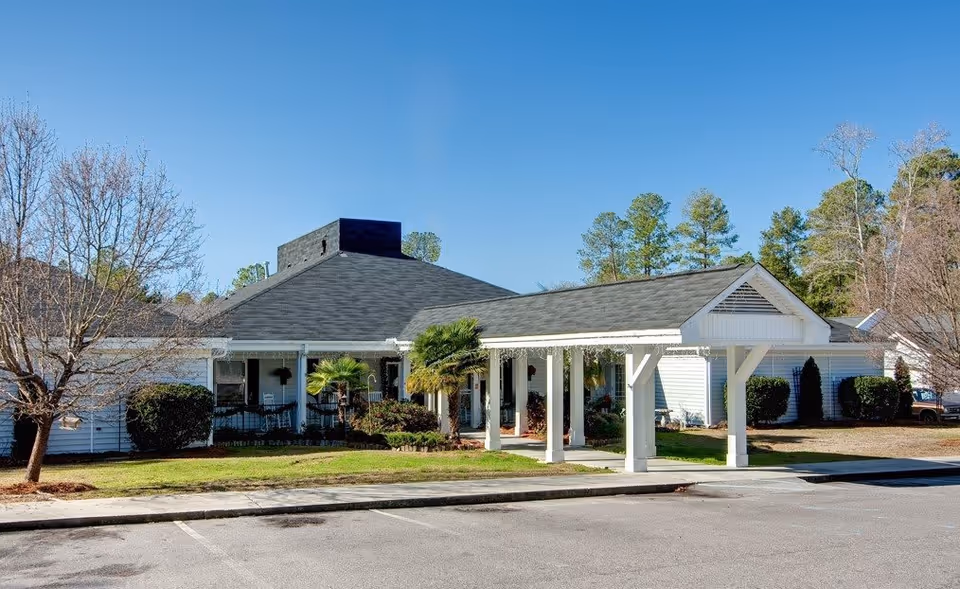 Exterior view of a single-story assisted living facility building with white siding, a covered entrance supported by white columns, and a well-maintained lawn with small trees and shrubs under a clear blue sky.