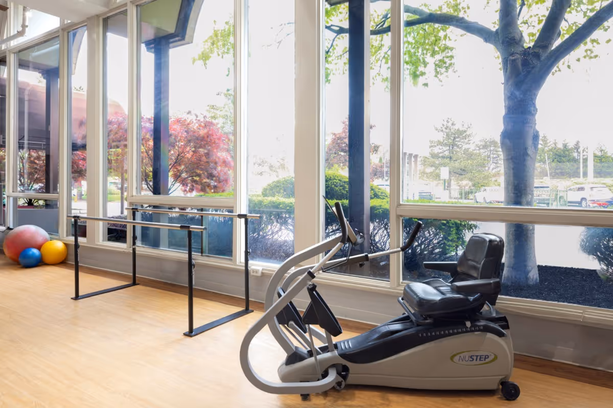A bright exercise room with large windows showing outdoor trees and parked cars. Inside, there is a recumbent exercise bike labeled NUSTEP, a ballet barre along the wall, and three colorful exercise balls in the corner on a wooden floor.