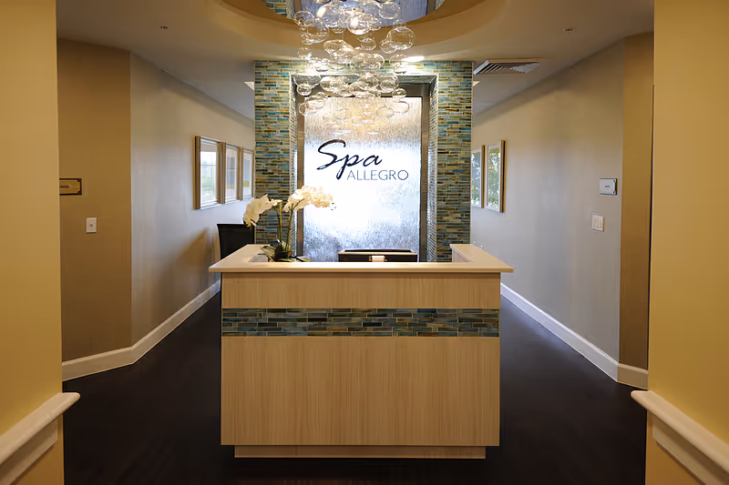 Reception desk area of Spa Allegro with a modern chandelier overhead, a decorative wall with the spa name, and a vase with white orchids on the desk. The space has warm lighting and neutral-colored walls with framed artwork.