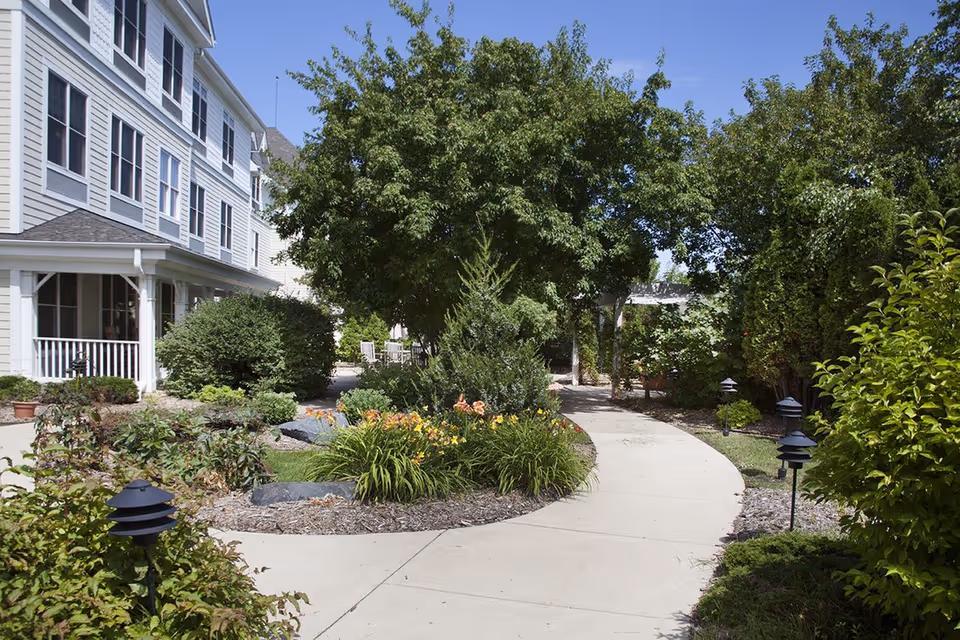 Curved walkway through a landscaped garden beside the exterior of a multi-story senior living building.