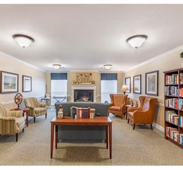 A cozy living room with a fireplace centered on the far wall between two windows with blue shades. The room contains a blue sofa facing away from the camera, a wooden table with books behind the sofa, two striped armchairs on the left, two brown leather armchairs on the right, a wooden bookshelf filled with books on the right side, and framed artwork on the walls. The room is well-lit with ceiling lights and a table lamp.