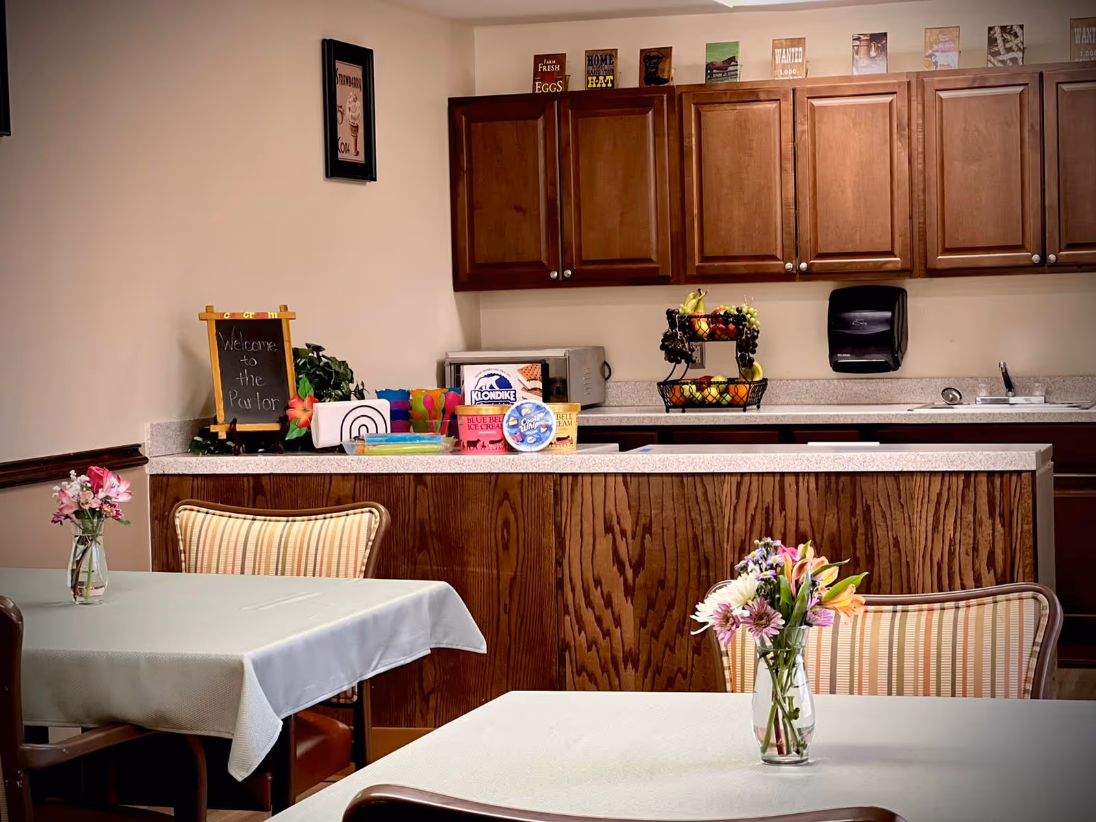 Interior view of a small dining area with tables covered in light-colored tablecloths and vases of flowers on each table. In the background, there is a kitchen counter with wooden cabinets above and various items including a fruit basket, ice cream containers, and a small chalkboard sign that reads 'Welcome to the Parlor'.