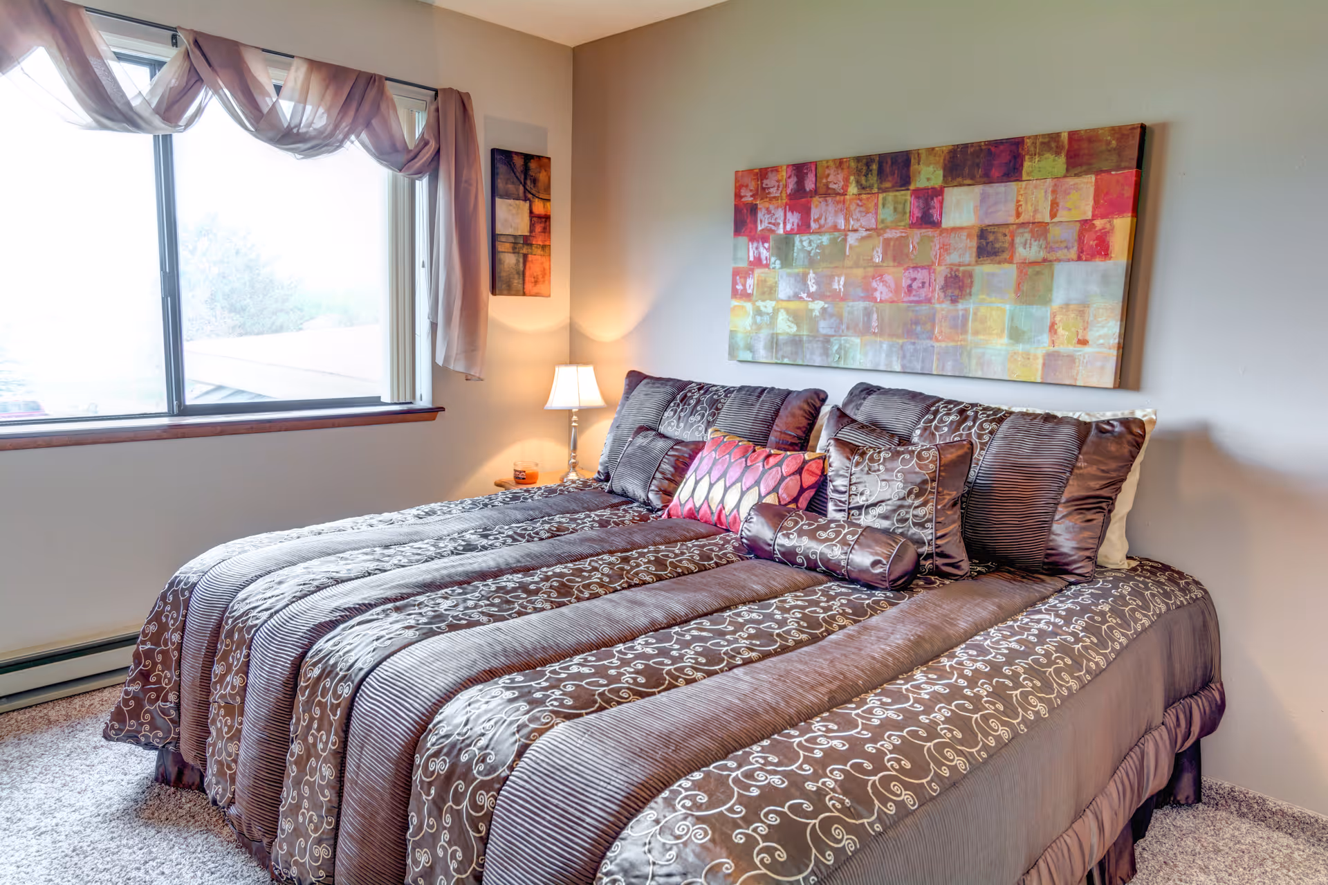 A neatly made bed with multiple decorative pillows and a patterned brown comforter in a bedroom. There is a large window with sheer curtains on the left side, a small bedside table with a lamp, and colorful abstract artwork hanging on the wall above the bed.