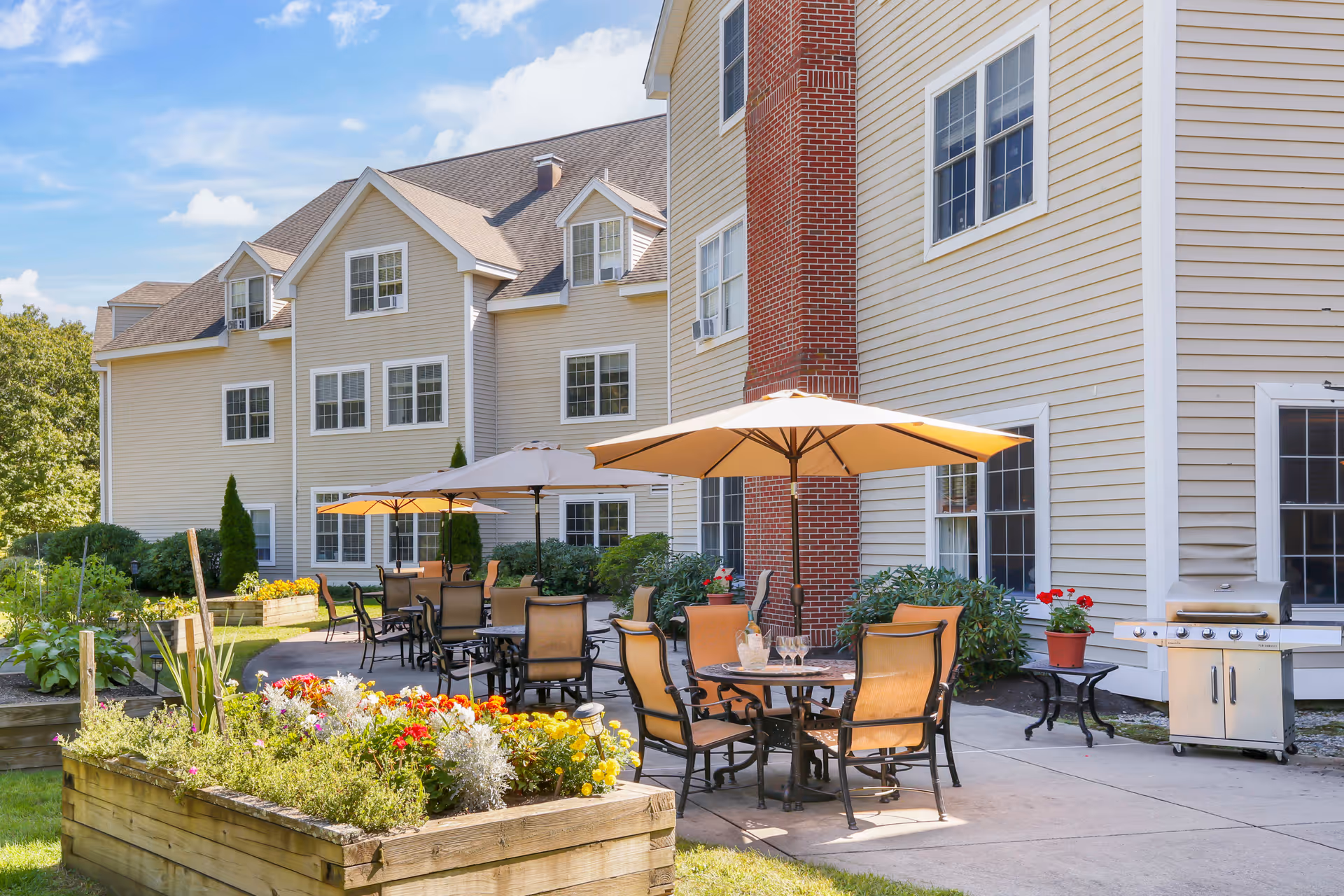 Outdoor patio area at Brookdale Attleboro featuring multiple tables with umbrellas and chairs, a barbecue grill, and raised garden beds with colorful flowers and greenery, adjacent to a beige multi-story building with white-framed windows and a red brick chimney under a partly cloudy blue sky.