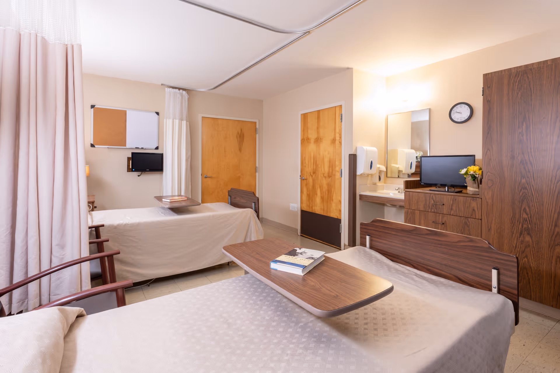 A well-lit room in a senior living facility with two hospital-style beds separated by a curtain. Each bed has a wooden overbed table, one with a book on it. There are wooden doors, a wall-mounted TV, a bulletin board, a clock, a sink with a mirror, and a wooden dresser with a small TV and a vase of flowers on top.