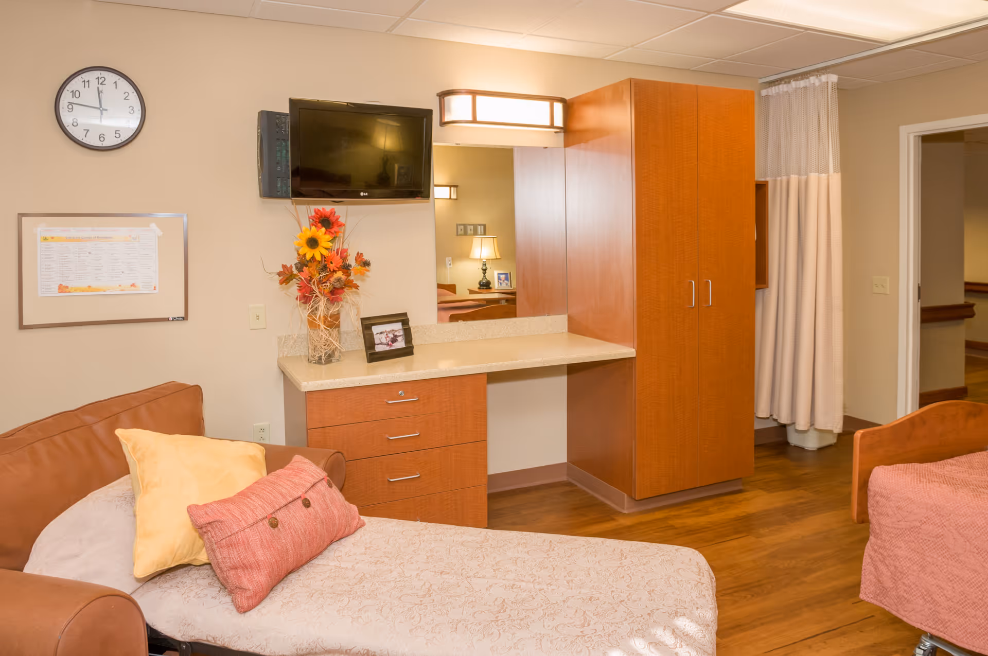 A cozy room in a senior living facility featuring a brown armchair with two pillows, a wooden cabinet with drawers and a countertop, a wall-mounted flat screen TV, a clock, a framed schedule, and a floral arrangement. The room has wooden flooring, a bed partially visible on the right, and a curtain dividing part of the space.