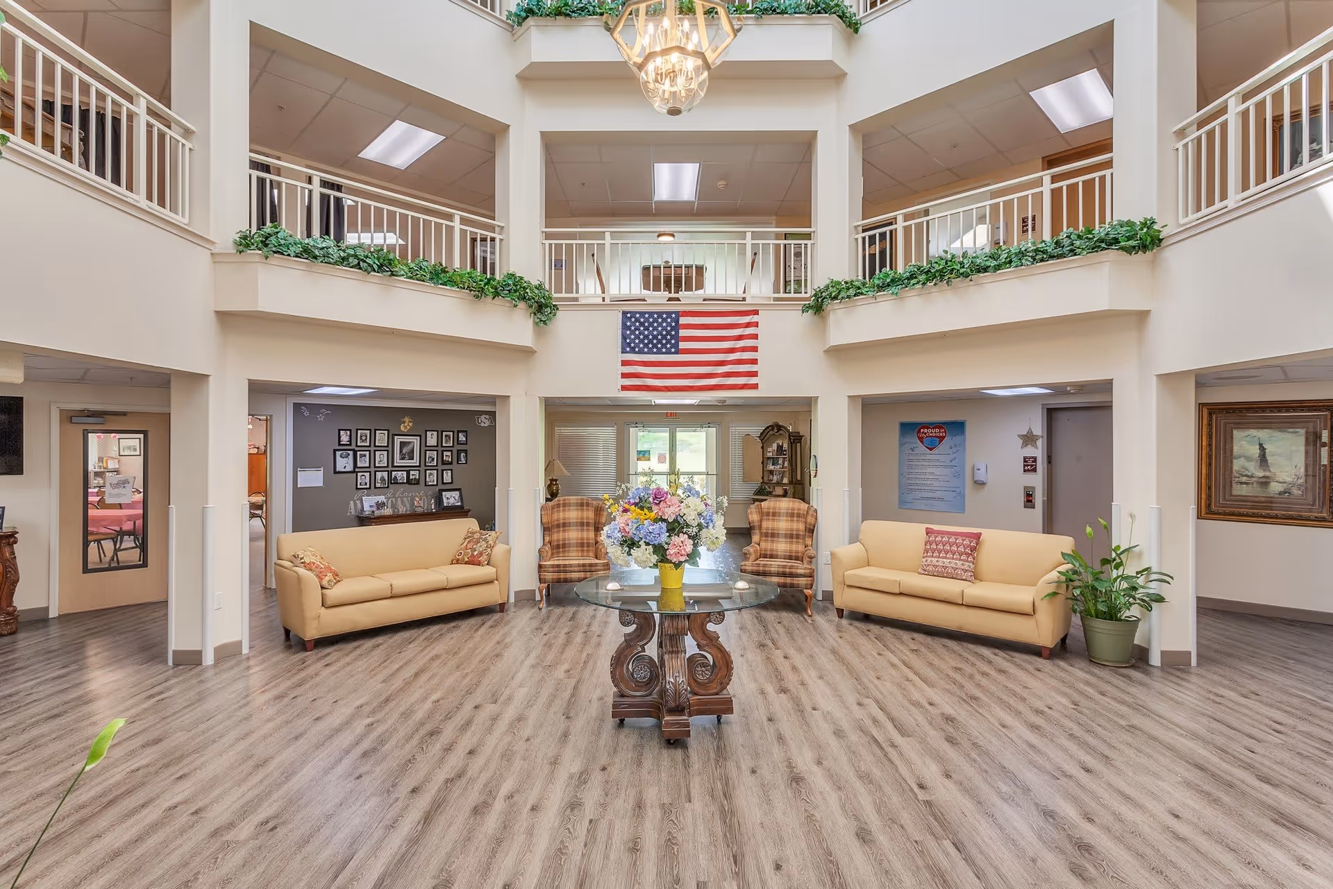 Spacious and well-lit common area in a senior living facility with two beige sofas, two plaid armchairs, a round glass table with a floral arrangement in the center, an American flag hanging on the wall, and green plants decorating the space. The area features wood flooring, a high ceiling with a chandelier, and a second-floor balcony with white railings.