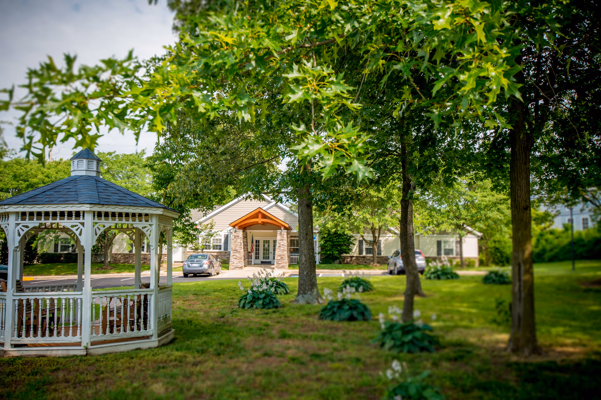 White gazebo on a grassy, tree-filled lawn in front of an assisted living building with a covered entrance and parked cars.