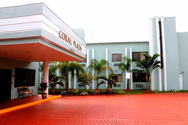 Exterior view of Coral Plaza senior living facility with turquoise walls, palm trees, and a driveway.