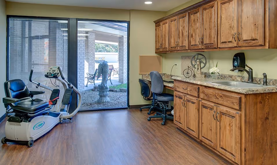 A room with wooden cabinets and a countertop with a sink on the right side, two office chairs at a desk area, and a recumbent exercise bike on the left side. A large glass door at the back shows an outdoor area with benches and a statue.