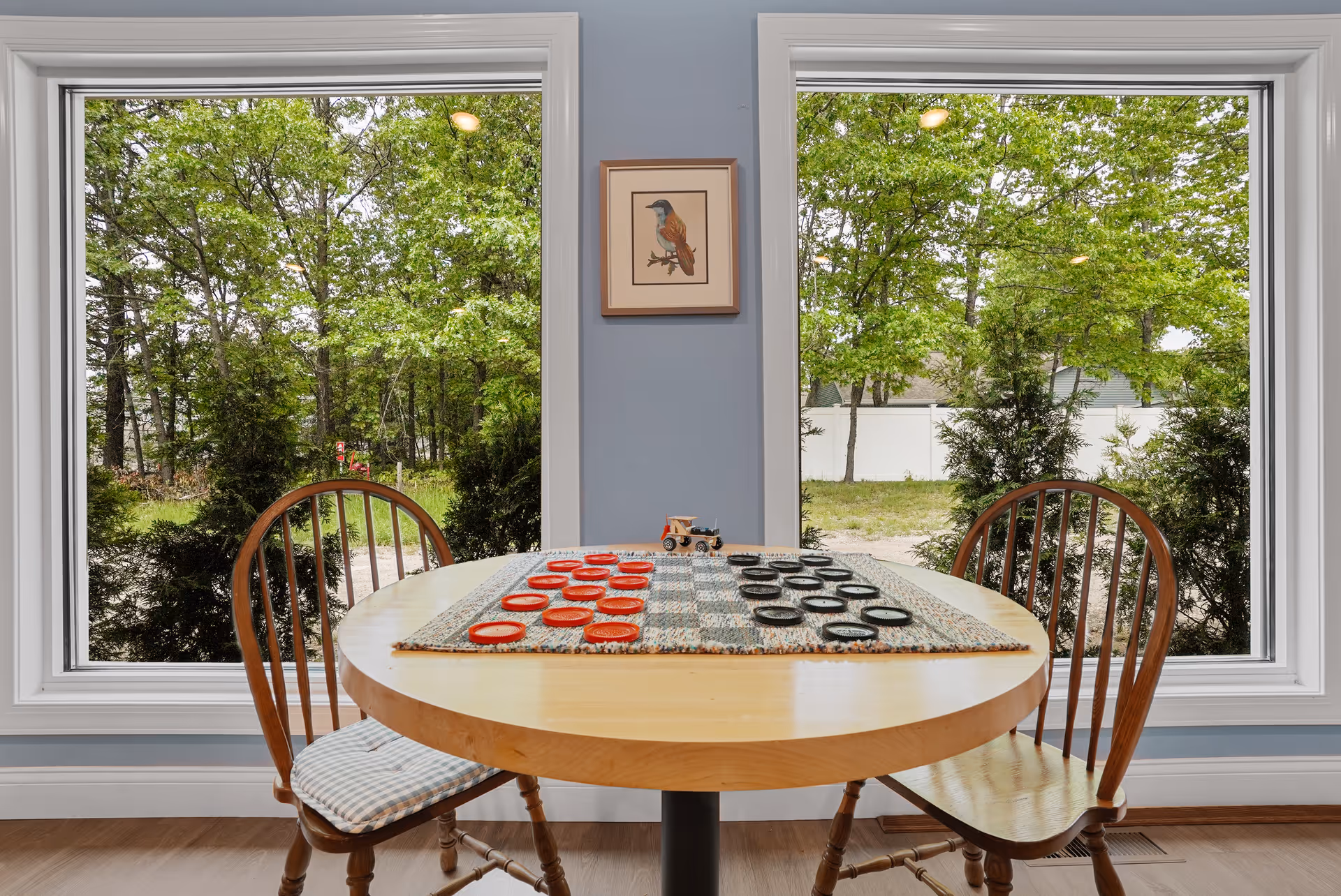 A round wooden table with a checkers game set up on a checkered board, positioned between two wooden chairs with cushions. The table is placed in front of two large windows showing green trees and a white fence outside. A framed picture of a bird is hung on the blue wall between the windows.