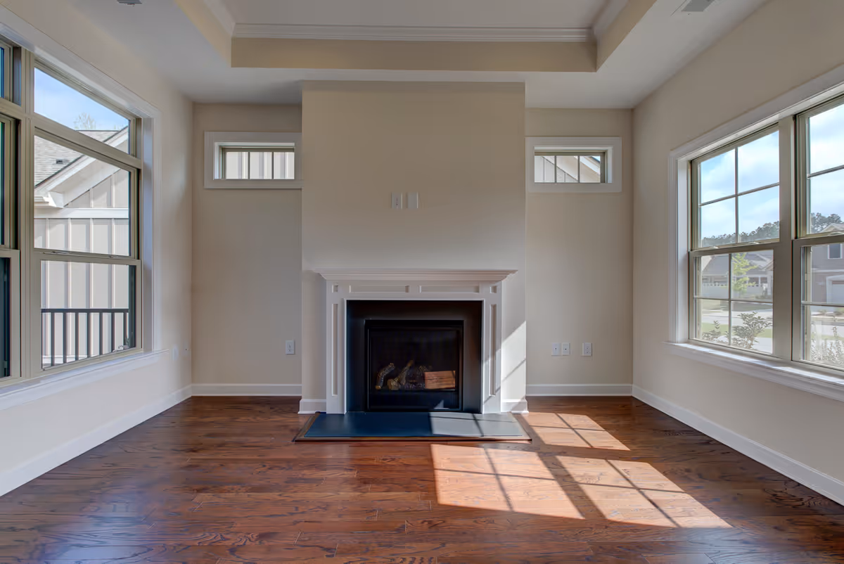 Empty room with hardwood floors, a white fireplace centered on the far wall, and large windows on both sides letting in natural light.