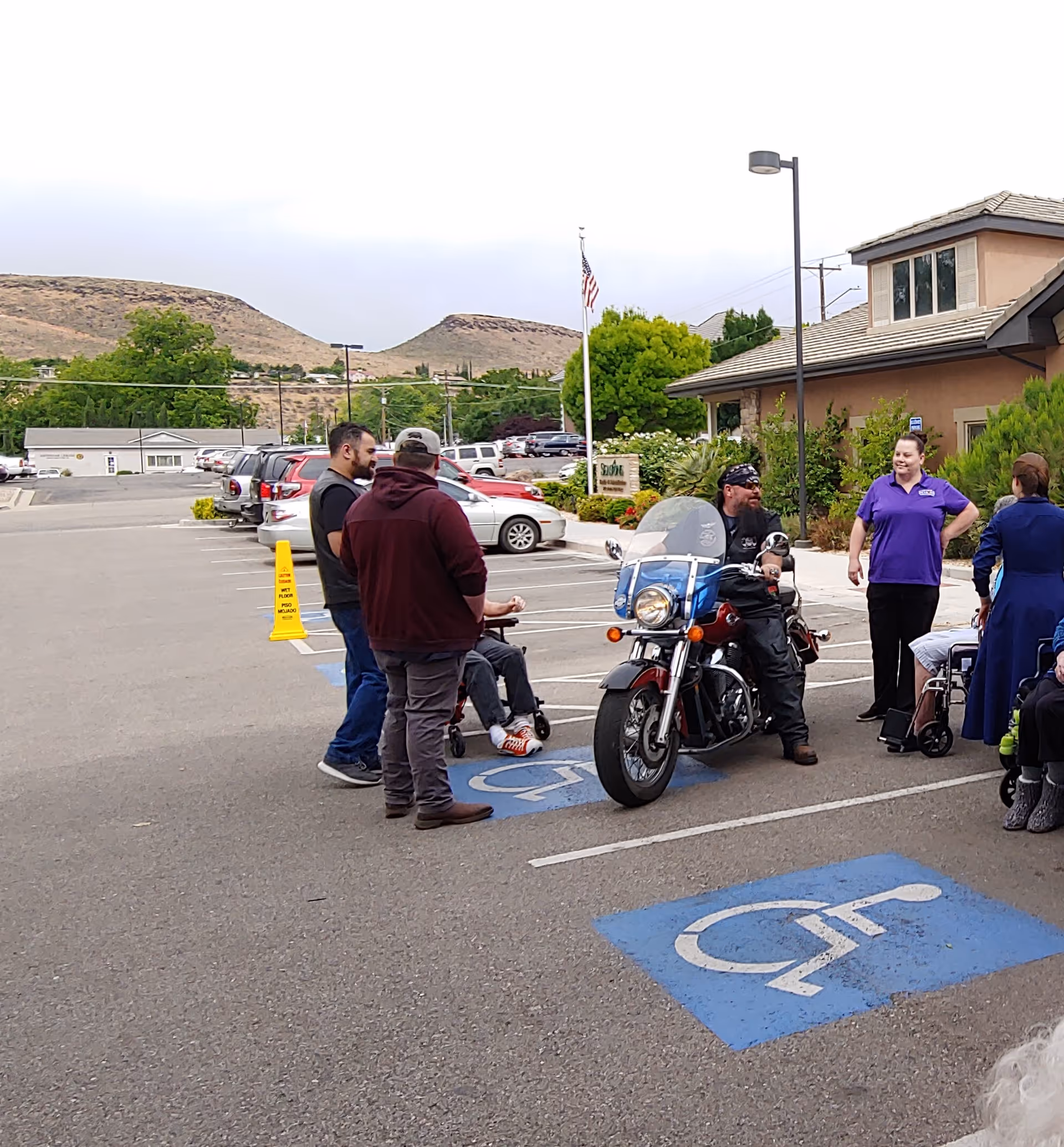 A group of people gathered in a parking lot outside a building, with one person sitting on a motorcycle. The parking lot includes marked handicapped parking spaces. In the background, there are parked cars, trees, and hills under a cloudy sky.