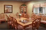 A dining room with a large wooden table surrounded by wooden chairs with cushioned seats. The table has a decorative inlay and a small flower vase in the center. The room features wallpapered walls, a framed painting, a glass-fronted corner cabinet with plants on top, and a window with floral valances.