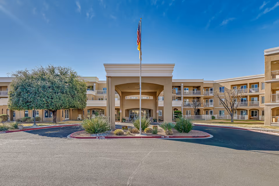 Front exterior view of Solstice Senior Living at Las Cruces building with a covered entrance, an American flag on a flagpole, landscaping with bushes and trees, and a clear blue sky.