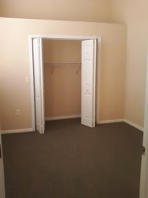 Empty room with beige walls and carpeted floor featuring an open closet with white bi-fold doors and a wire shelf inside.