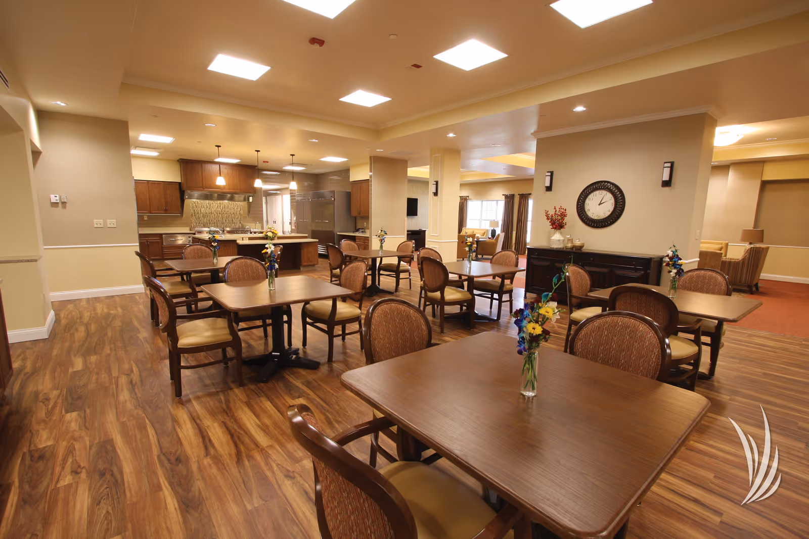 A spacious dining area with several wooden tables and cushioned chairs arranged neatly. Each table has a small vase with colorful flowers. The room features wood flooring, warm beige walls, and ceiling lights. In the background, there is a kitchen area with wooden cabinets, a large refrigerator, and pendant lights hanging over a kitchen island. A clock and decorative items are on a sideboard against one wall.