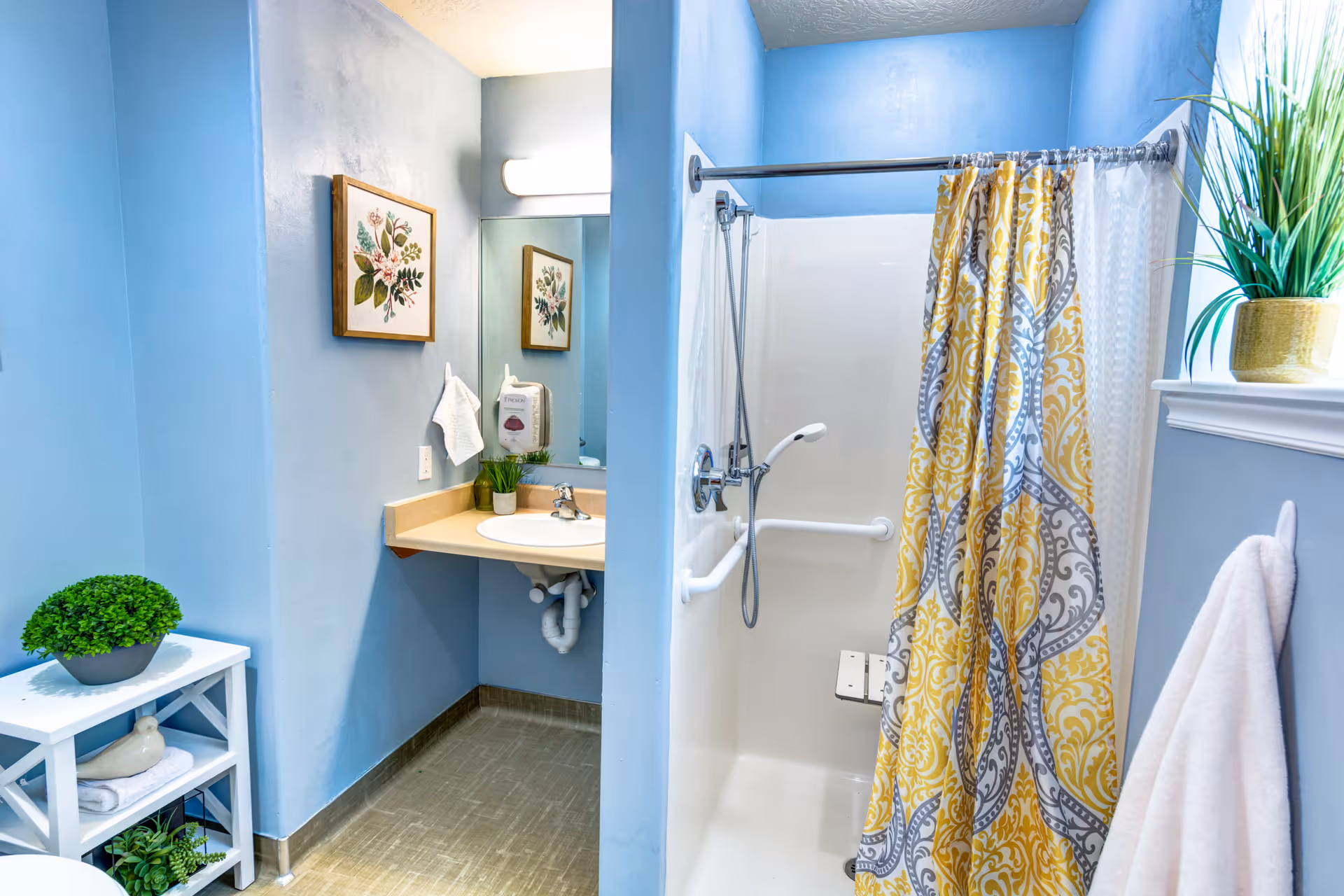 Bright blue-painted bathroom with a walk-in shower and yellow patterned curtain, a sink vanity with mirror, and decorative plants and towels.