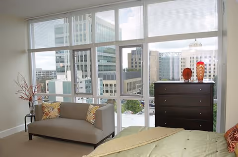 A bright bedroom with large floor-to-ceiling windows showing a cityscape view. The room contains a beige sofa with patterned cushions, a dark wooden dresser with decorative vases on top, and a bed partially visible with a green comforter and a red patterned pillow.