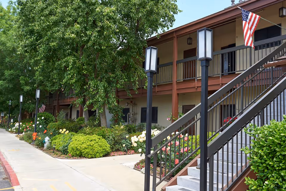 Exterior view of a two-story residential building with a staircase leading to the upper floor. The building is surrounded by lush greenery, bushes, and colorful flowers. Several lamp posts line the sidewalk, and an American flag is displayed near the staircase.