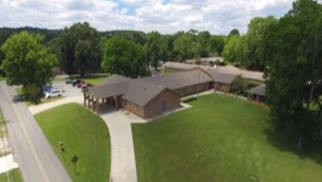 Aerial view of Burns Nursing Home showing a single-story building with a brown roof surrounded by a large green lawn and trees. A driveway leads to the entrance, and several cars are parked near the building. The sky is partly cloudy.