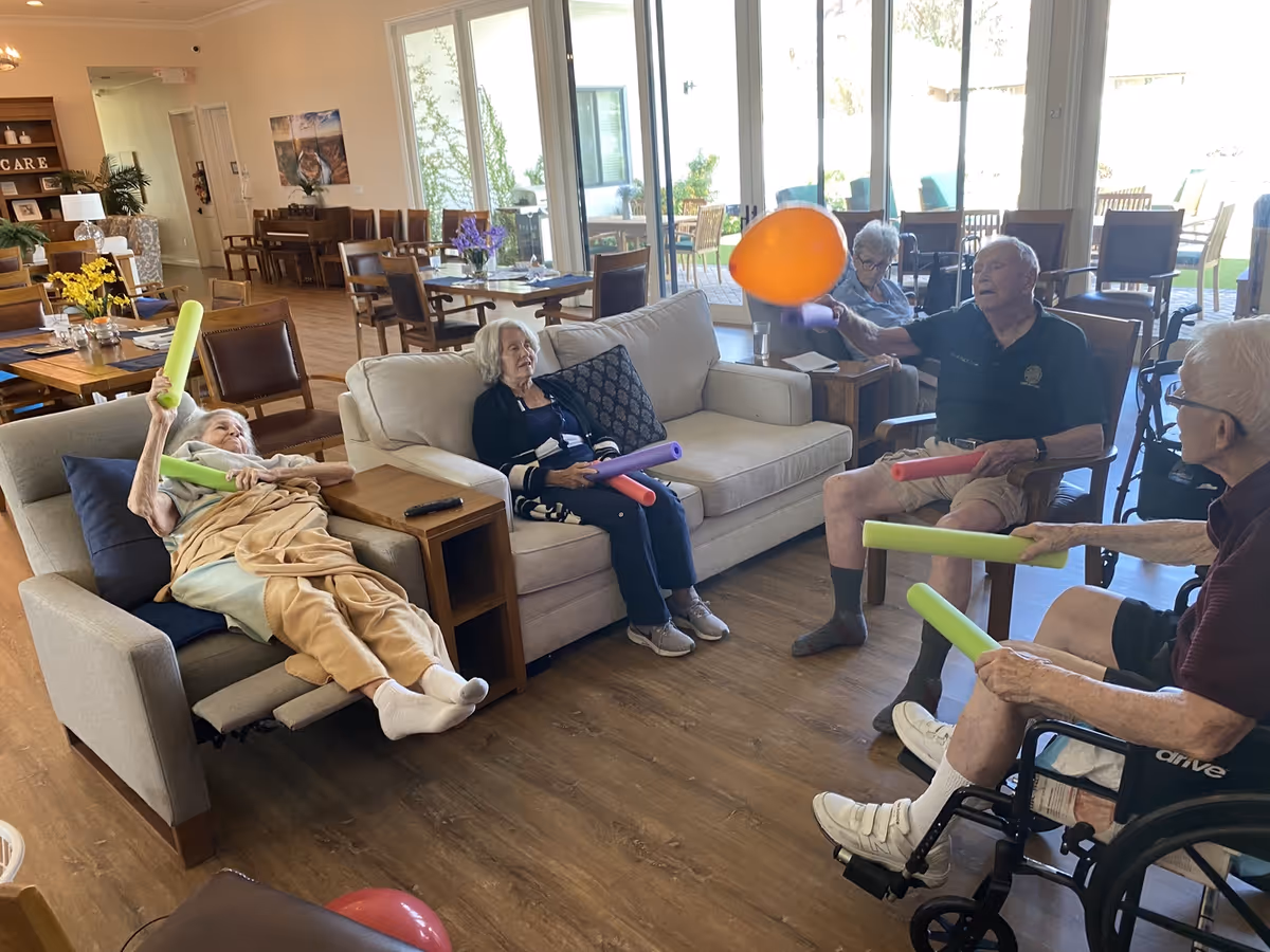 Elderly residents in a senior living lounge sitting in chairs and a recliner playing with pool noodles and an orange balloon.