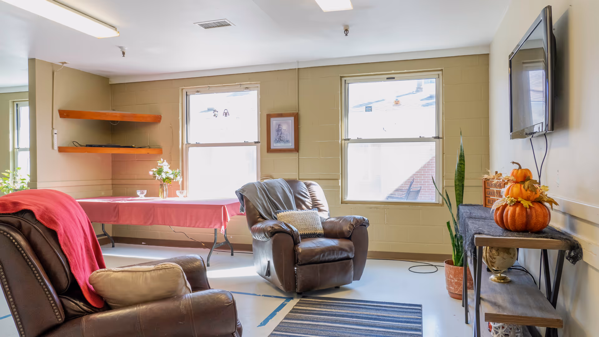 A cozy living room area with two brown leather armchairs, one draped with a red blanket and the other with a gray throw and a decorative pillow. There is a striped rug on the floor, a table with a red tablecloth near two windows, and a wall-mounted TV above a console table decorated with pumpkins and a potted plant. The room has beige walls and large windows letting in natural light.