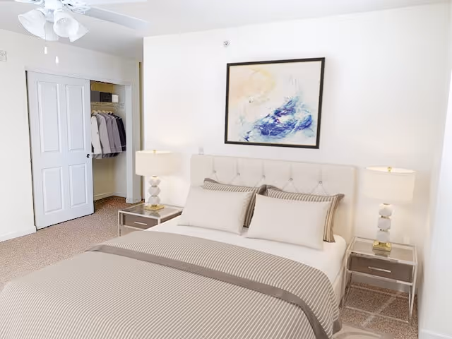 A bright and clean bedroom with a neatly made bed featuring beige and white striped bedding. There are two matching bedside tables with modern lamps on each side of the bed. A framed abstract painting hangs on the wall above the headboard. An open closet with hanging clothes is visible to the left.