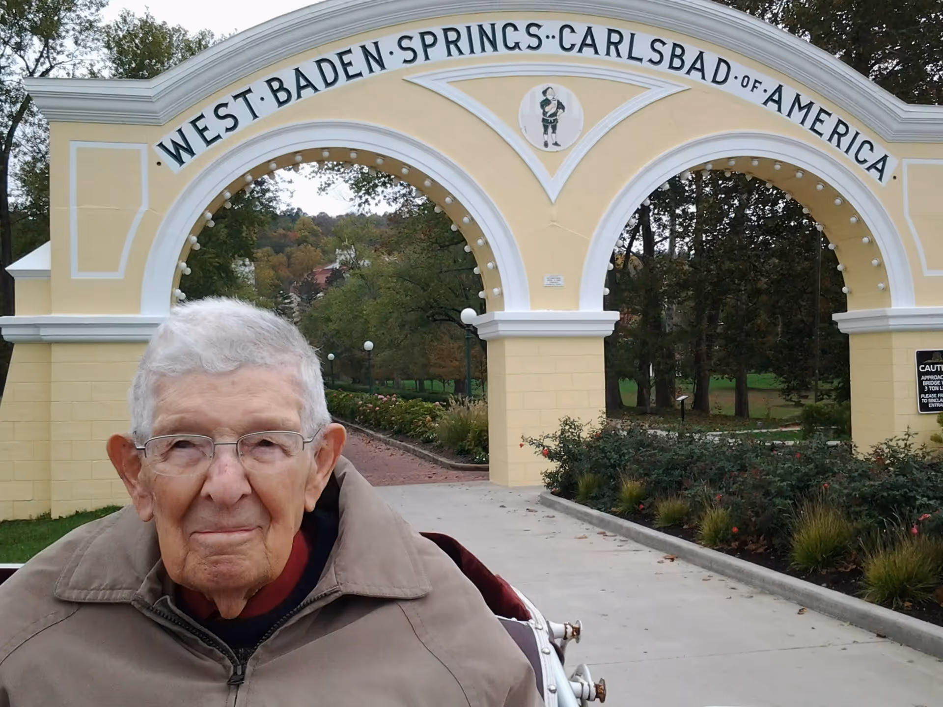 A person seated in a wheelchair in front of a pale yellow decorative archway reading 'WEST BADEN SPRINGS · CARLSBAD OF AMERICA'.