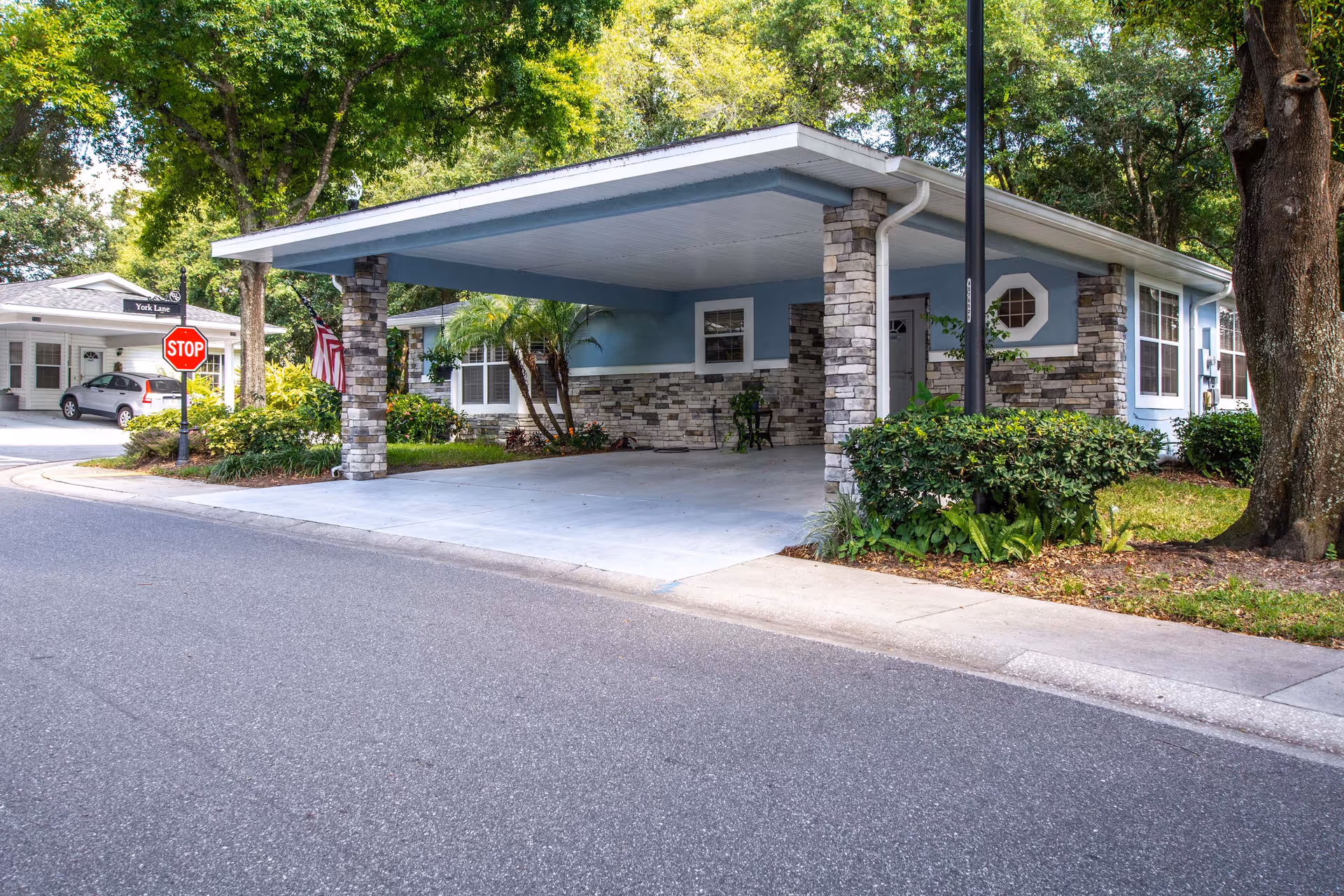 Exterior view of a single-story residential building with a covered carport supported by stone pillars. The building has light blue siding with stone accents and white trim around the windows. There is a stop sign and a street sign labeled York Lane near the driveway, surrounded by greenery and trees.
