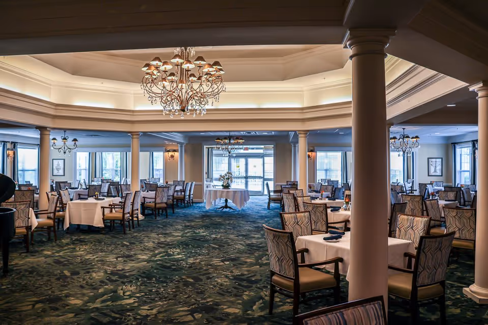 Spacious dining room in a senior living facility with multiple tables covered in white tablecloths and surrounded by patterned chairs. The room features large windows allowing natural light, decorative chandeliers hanging from the ceiling, and columns supporting the ceiling structure.