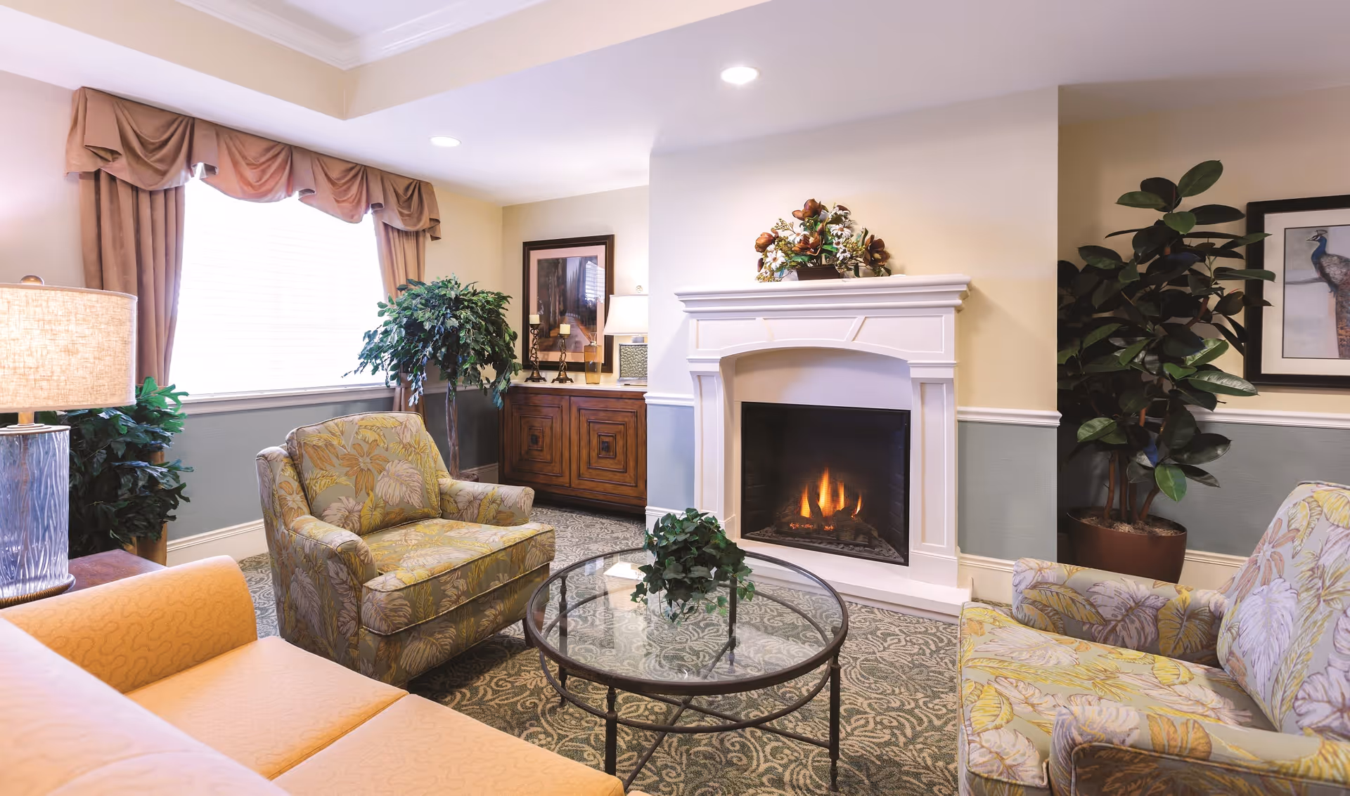 A cozy living room with a lit fireplace, two patterned armchairs, a peach-colored sofa, a glass coffee table with a small plant, a wooden cabinet with a lamp and framed artwork, large windows with drapes, and several potted plants.