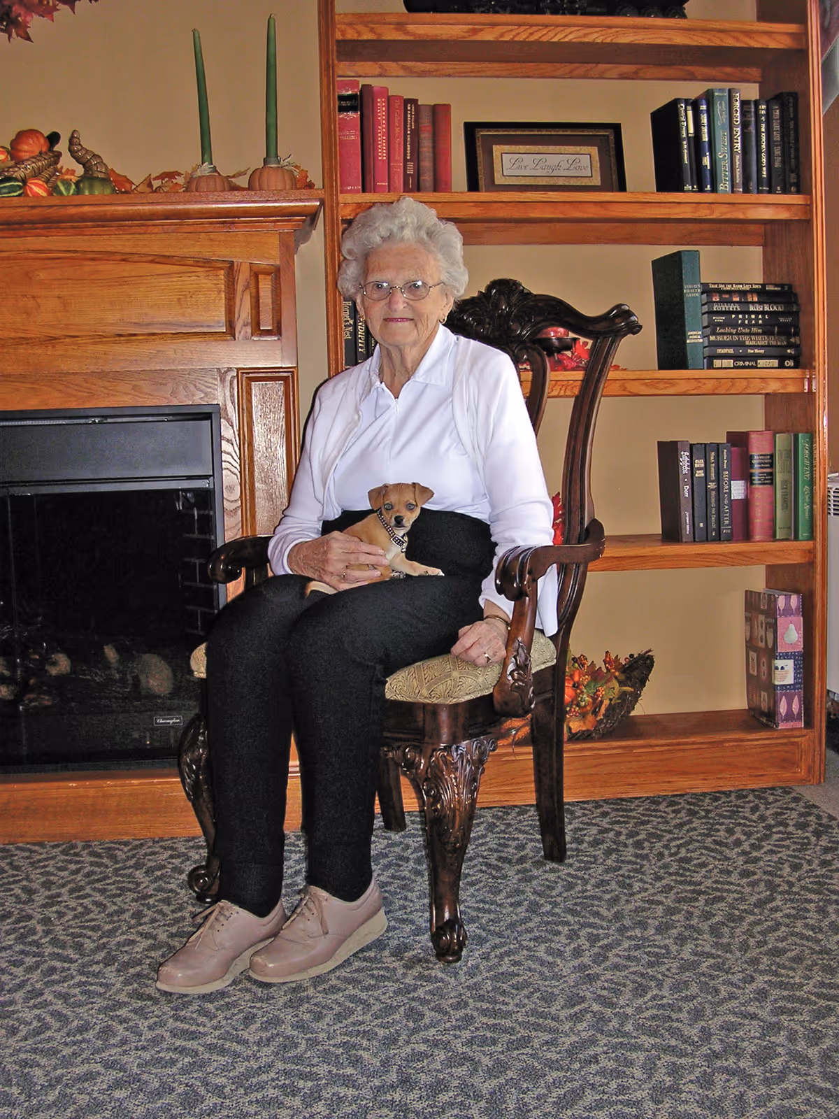 An elderly woman with white hair and glasses sitting on an ornate wooden chair in a room with a fireplace and wooden bookshelves. She is wearing a white long-sleeve shirt and black pants, holding a small brown dog on her lap. The bookshelves behind her contain books and decorative items.