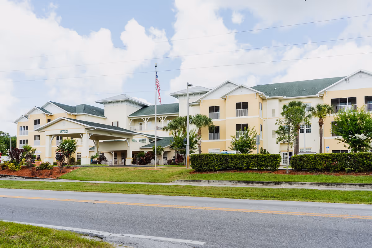 Exterior view of a large, three-story senior living facility building with a covered entrance, manicured landscaping, palm trees, and an American flag on a flagpole in front. The building has a beige and white facade with green roofing under a partly cloudy sky.