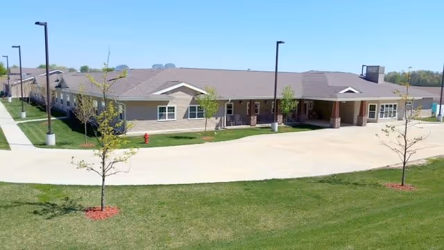 Exterior view of a single-story assisted living facility building with a large driveway and small trees planted in front on a grassy lawn under a clear sky.