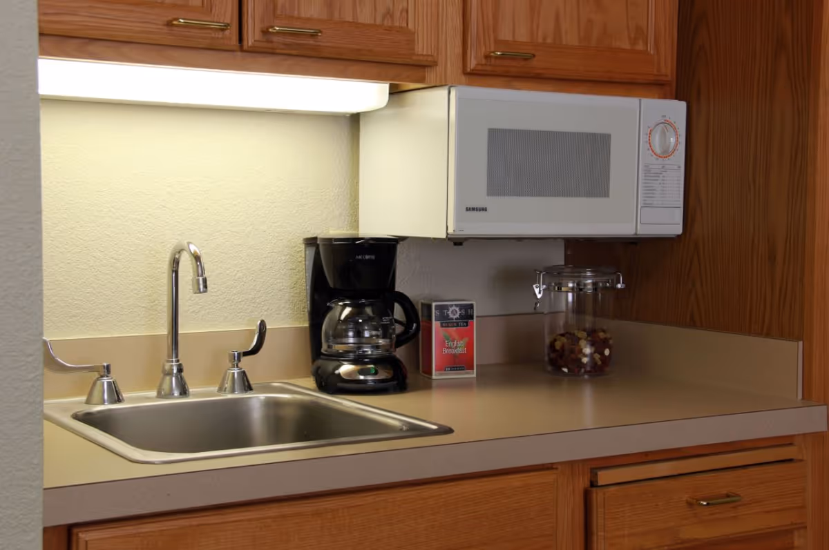 A kitchen countertop with a stainless steel sink, a coffee maker, a box of English Breakfast tea, a glass jar with a metal clasp containing dried fruit or nuts, and a white microwave mounted above the counter. Wooden cabinets are visible above and below the countertop.