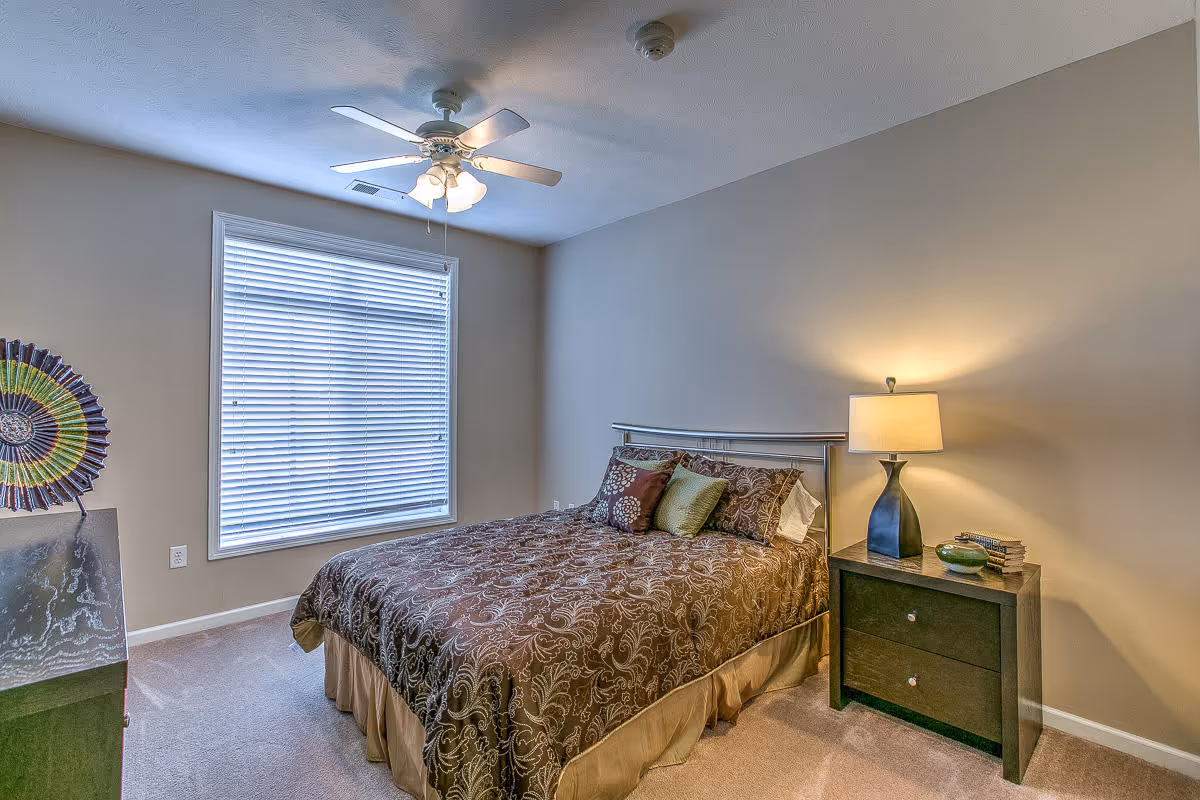 A bedroom with a large window covered by white blinds, a ceiling fan with lights, a bed with a brown patterned comforter and several pillows, a nightstand with a lamp, books, and a decorative bowl, and a piece of colorful wall art partially visible on the left.