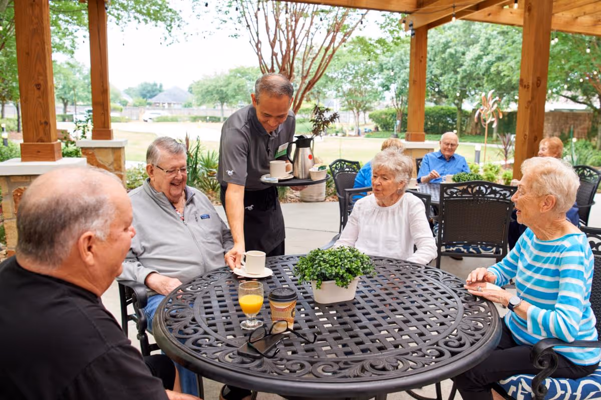 A group of elderly people sitting around a round outdoor metal table under a wooden pergola. A server is placing a cup on the table. The table has a small potted plant, a glass of orange juice, a coffee cup, and a pair of glasses. Trees and greenery are visible in the background.