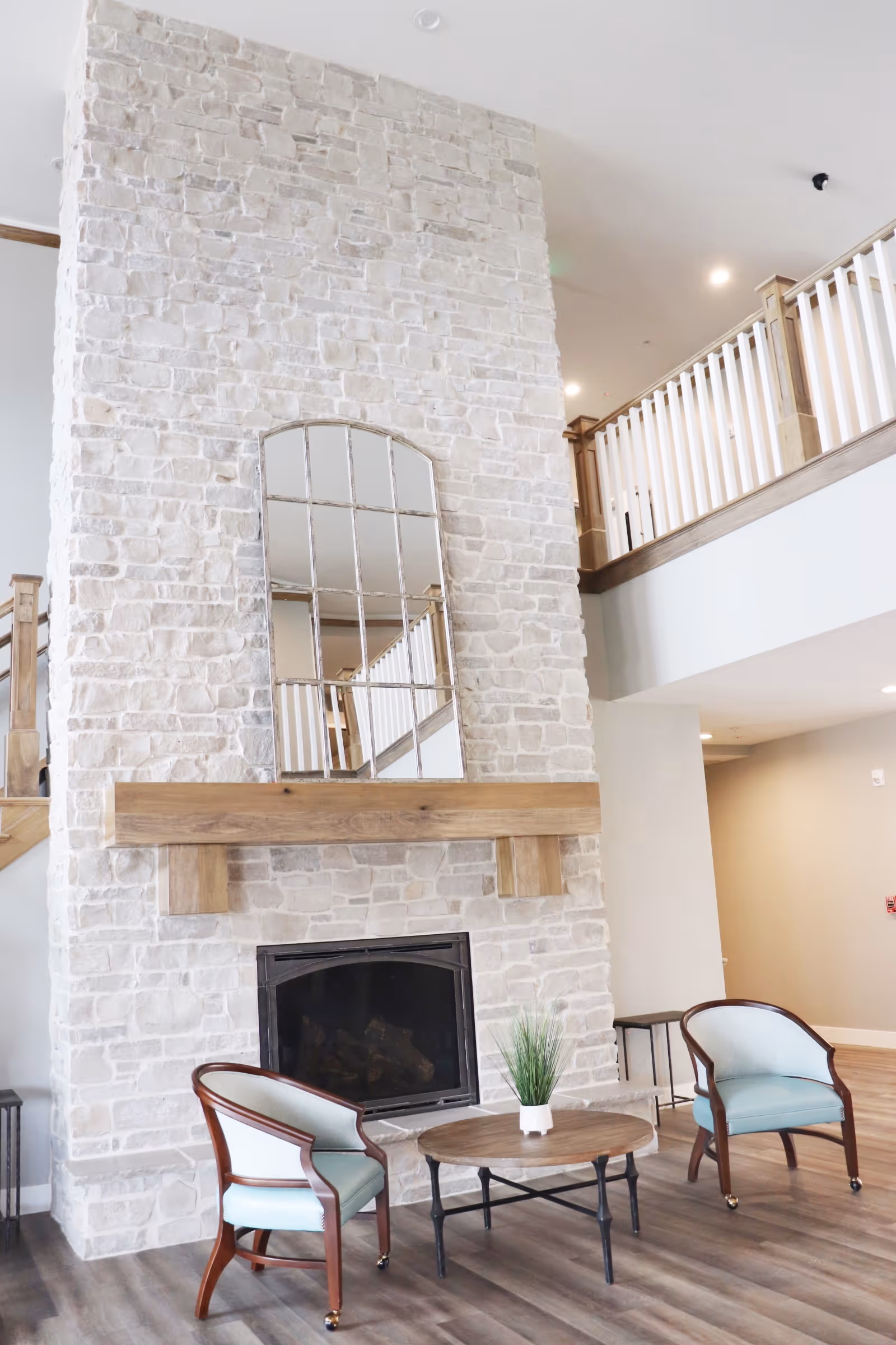 Spacious common lounge with a tall stone fireplace, mirror above the wooden mantel, two upholstered armchairs and a round coffee table.