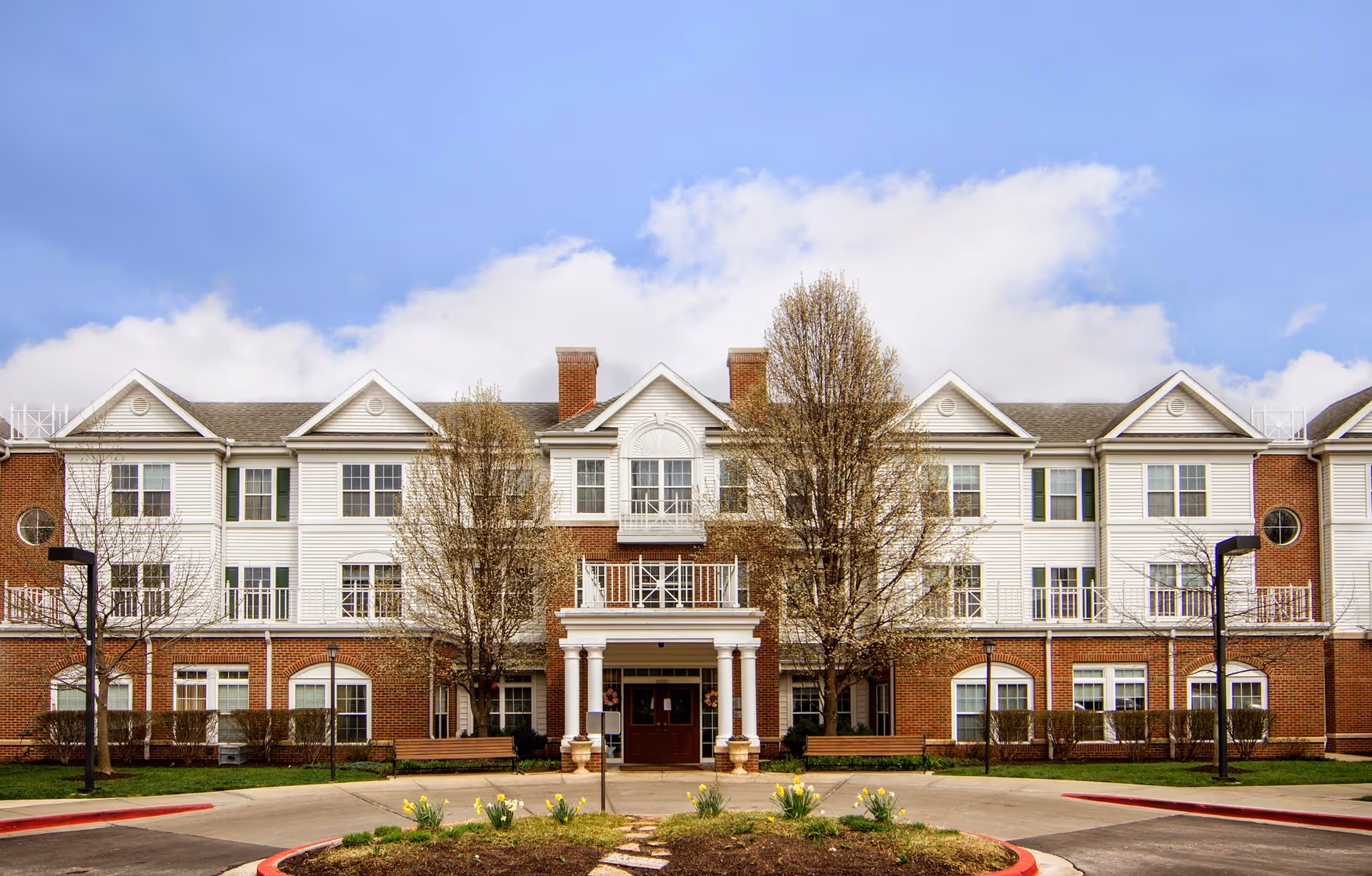 Front exterior view of a three-story senior living facility building with a combination of red brick and white siding. The entrance has a covered porch with white columns, and there are leafless trees and landscaped flower beds in front. The sky is partly cloudy.