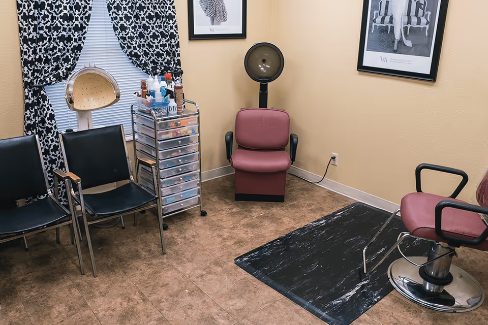 Interior of a small salon room with two black chairs, a rolling cart with various hair and beauty products, a maroon salon chair under a hair dryer, and another maroon salon chair with a footrest. The walls are beige with two framed black and white pictures, and there is a window with black and white patterned curtains.