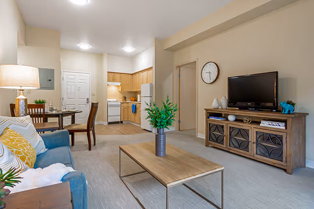 A bright and cozy living room area with a blue sofa adorned with patterned cushions, a wooden coffee table with a vase of green plants, a wooden TV stand with a flat-screen TV and decorative items, and a dining table with chairs near the kitchen. The kitchen features wooden cabinets, a white refrigerator, and a stove. The walls are painted beige, and the room is well-lit with ceiling lights.