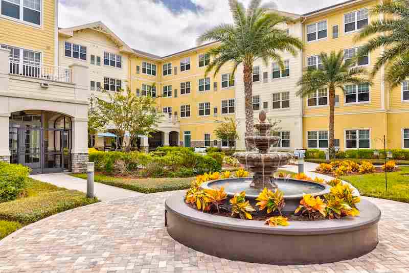 Outdoor courtyard area of a senior living facility with a multi-tiered water fountain surrounded by yellow and orange flowers. The courtyard is paved with bricks and features palm trees, shrubs, and a large yellow building with many windows in the background.
