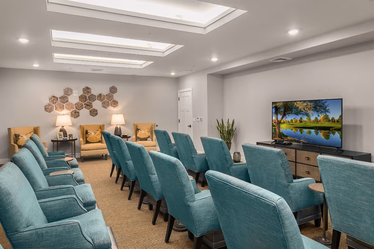 A bright common room with rows of teal armchairs facing a television, plus accent chairs, lamps, and wall decor.