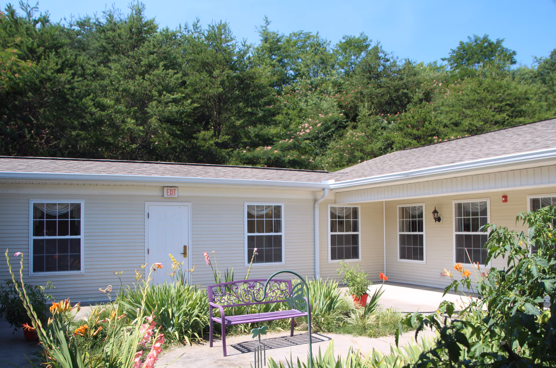 Small courtyard with a purple bench and flower beds in front of a single-story beige building and wooded hillside behind it.