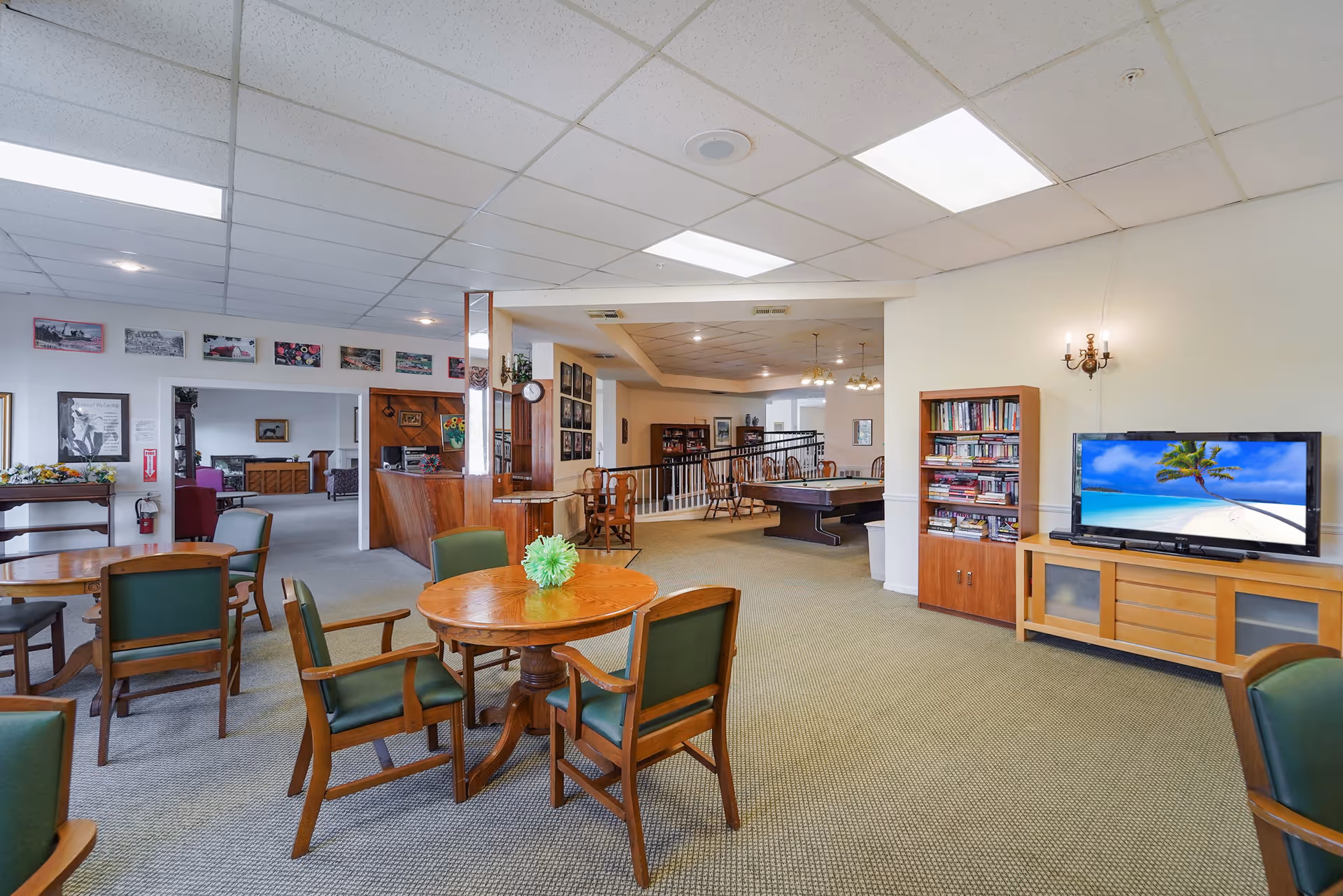 A spacious common area in an assisted living residence featuring several wooden tables with green cushioned chairs, a pool table, a bookshelf filled with books, and a television displaying a beach scene. The room has a carpeted floor, white walls, and a drop ceiling with fluorescent lighting. There are framed pictures on the walls and a small reception desk in the background.