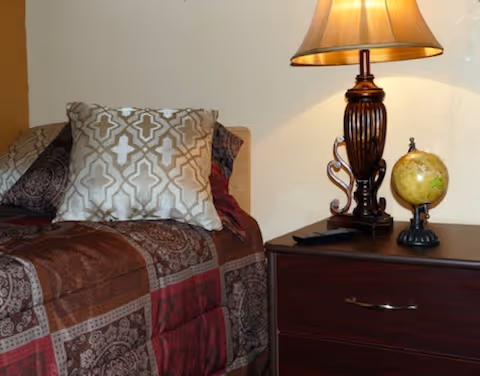 A cozy bedroom corner featuring a bed with patterned pillows and a quilted bedspread in shades of brown and red. Next to the bed is a dark wooden nightstand with a decorative table lamp and a small globe on it.