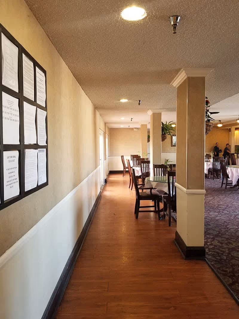 Interior view of a dining area in a senior living facility with wooden floors, beige walls, and several tables with chairs. The room is lit by ceiling lights, and there are plants hanging from the ceiling. Two people are visible in the background near the dining tables. A bulletin board with papers is mounted on the left wall.