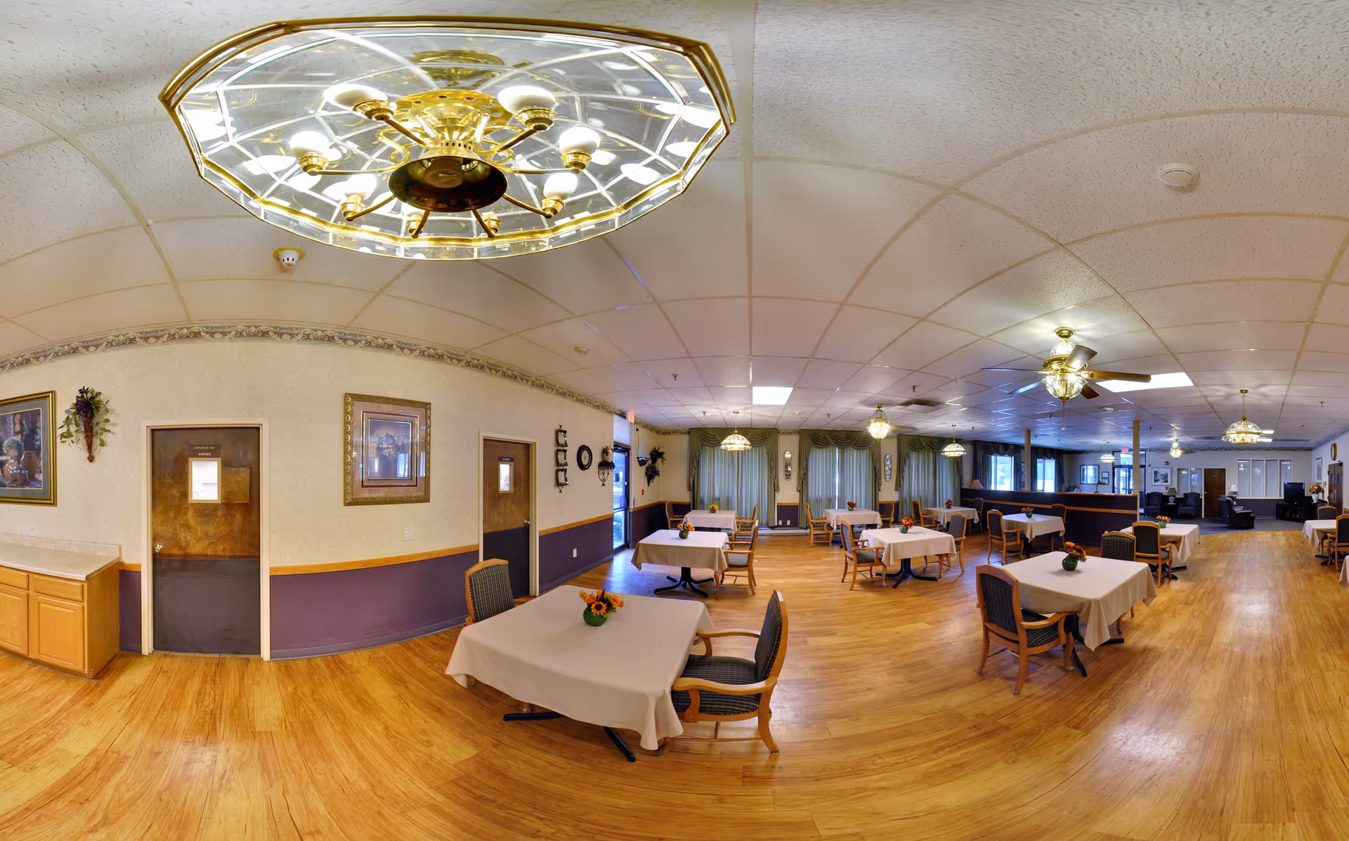 A spacious dining room with multiple tables covered in white tablecloths, each adorned with a small floral centerpiece. The room features wooden flooring, several ceiling fans with lights, and large windows with green curtains allowing natural light to enter. The walls are decorated with framed pictures and wall sconces. There are chairs neatly arranged around the tables, and the overall atmosphere is warm and inviting.